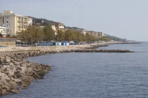 Una fotografia reale e panoramica scattata dalla costa che offre una vista suggestiva del mare verso l'insediamento urbano interessato dal caso burocratico; in primo piano le acque calme del litorale riflettono la luce del giorno, mentre sullo sfondo si estende il profilo del quartiere abusivo che si sviluppa lungo la costa con le sue abitazioni fitte e stratificate, prive di una pianificazione urbanistica ufficiale e di numeri civici visibili, evidenziando il contrasto tra la bellezza paesaggistica naturale e la complessa realtà sociale e amministrativa dei residenti.