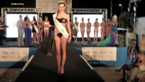 Fotografia della vincitrice Giulia Zancato durante la proclamazione sul palco della sesta edizione di Miss Venice Beach 2016. Indossa un costume nero, la fascia del titolo, la corona e il numero identificativo 10. Sorride mentre tiene in mano alcune borse premio. Sullo sfondo sono presenti altre concorrenti in costume e fascia, la passerella illuminata, pannelli con loghi degli sponsor e luci da palco. L’immagine cattura il momento della vittoria e della consegna dei premi.