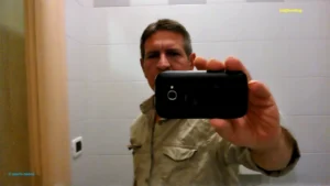 Immagine verticale che mostra Mario mentre si scatta una foto allo specchio con uno smartphone, in un ambiente con piastrelle chiare e una porta in legno sul lato. Indossa una camicia beige e tiene il telefono davanti al volto, con le scritte “© Mario Bonesi” e “Gidiferroblog” visibili nell’immagine.