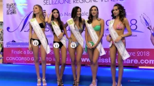 Fotografia scattata sul palco della selezione regionale Veneto di Miss Blumare. Sono presenti le concorrenti fasciate, in costume da passerella, disposte in linea davanti al banner ufficiale dell’evento. Da sinistra: Pamela Grotto, Irene Cappelletti, Alessia Alberti, Giada Da Rugna e Alessia Peron, tutte con fascia identificativa e numero di gara. Il fondale riporta la grafica “Selezione Regione Veneto” e loghi del concorso. L’illuminazione è uniforme e mette in risalto le fasce e la postura delle partecipanti.