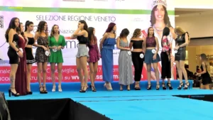 Fotografia delle concorrenti di Miss Blumare Veneto mentre effettuano le prove sul palco prima della selezione ufficiale. Le ragazze indossano abiti casual o da passerella, con numeri di gara visibili, e seguono le indicazioni dello staff. Sullo sfondo si nota il banner “Selezione Regione Veneto” e la grafica del concorso nazionale. L’atmosfera è operativa e preparatoria, con illuminazione da palco e pubblico che osserva dalle aree circostanti del centro commerciale.