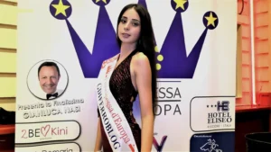 Foto di una  concorrente, Valeria Nicoli, indossa una corona, la fascia con le diciture Vogue, Principessa d’Europa e Miss Triveneto, e tiene un bouquet di fiori. È posizionata davanti al pannello ufficiale dell’evento, decorato con una grande corona stilizzata e loghi degli sponsor, tra cui Hotel Eliseo Riccione, Paolo DeMaria make‑up artist e Gidiferroblog. L’illuminazione mette in risalto la postura, la fascia e i dettagli del costume da passerella.