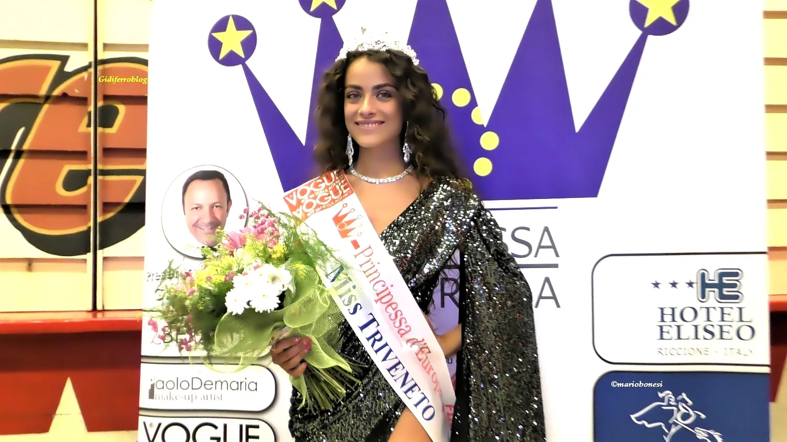 Fotografia della vincitrice Aurora Maglione, della selezione Triveneto di Miss Principessa d’Europa. La concorrente indossa una corona, la fascia con le diciture Vogue, Principessa d’Europa e Miss Triveneto, e tiene un bouquet di fiori. È posizionata davanti al pannello ufficiale dell’evento, decorato con una grande corona stilizzata e loghi degli sponsor, tra cui Hotel Eliseo Riccione, Paolo DeMaria make‑up artist. L’illuminazione mette in risalto la postura, la fascia e i dettagli del costume da passerella.