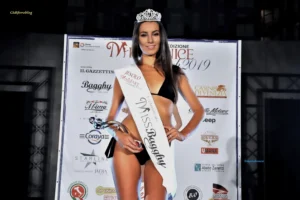 Fotografia di Valentina Pecchi, vincitrice della selezione Miss Venice Beach 2019 a Jesolo. Indossa un bikini nero, la corona e la fascia Jesolo Miss Venice Beach 2019 – 1ª Classificata Miss Bagghy, e tiene in mano il trofeo della serata. Sullo sfondo è visibile il pannello ufficiale del concorso con loghi degli sponsor, tra cui Il Gazzettino, Bagghy, Casinò di Venezia, Coraya, Starline e Umana. L’immagine documenta il momento della proclamazione della vincitrice.