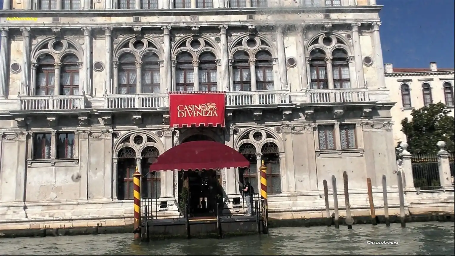 Fotografia dell’ingresso del Casinò di Venezia ripreso dal Canal Grande. L’edificio storico presenta una facciata rinascimentale in pietra chiara con finestre ad arco, colonne decorative e dettagli scolpiti. In primo piano si vede il pontile d’accesso con pali veneziani a righe bianche e blu, mentre sopra l’ingresso è collocato un grande telo rosso con la scritta “Casinò di Venezia”. L’immagine documenta l’accesso via acqua alla sede della finale di Miss Venice Beach.