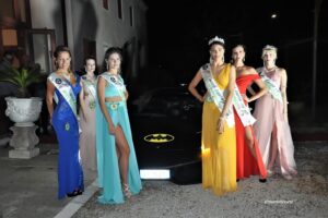 Una fotografia di gruppo che ritrae le sei ragazze premiate della selezione di Miss Earth Italy 2019 in Veneto. Le miss posano sorridenti ed eleganti disposte attorno a un'automobile posizionata scenograficamente accanto al palco. Tutte le concorrenti indossano gli abiti da sera della finale e le fasce ufficiali del concorso, inclusi i titoli legati agli elementi naturali. L'immagine combina il fascino delle protagoniste con lo stile della vettura, creando uno scatto promozionale classico dei concorsi di bellezza all'aperto. Maria Teresa Gonzalez risalta al centro del gruppo con la corona di vincitrice della serata.