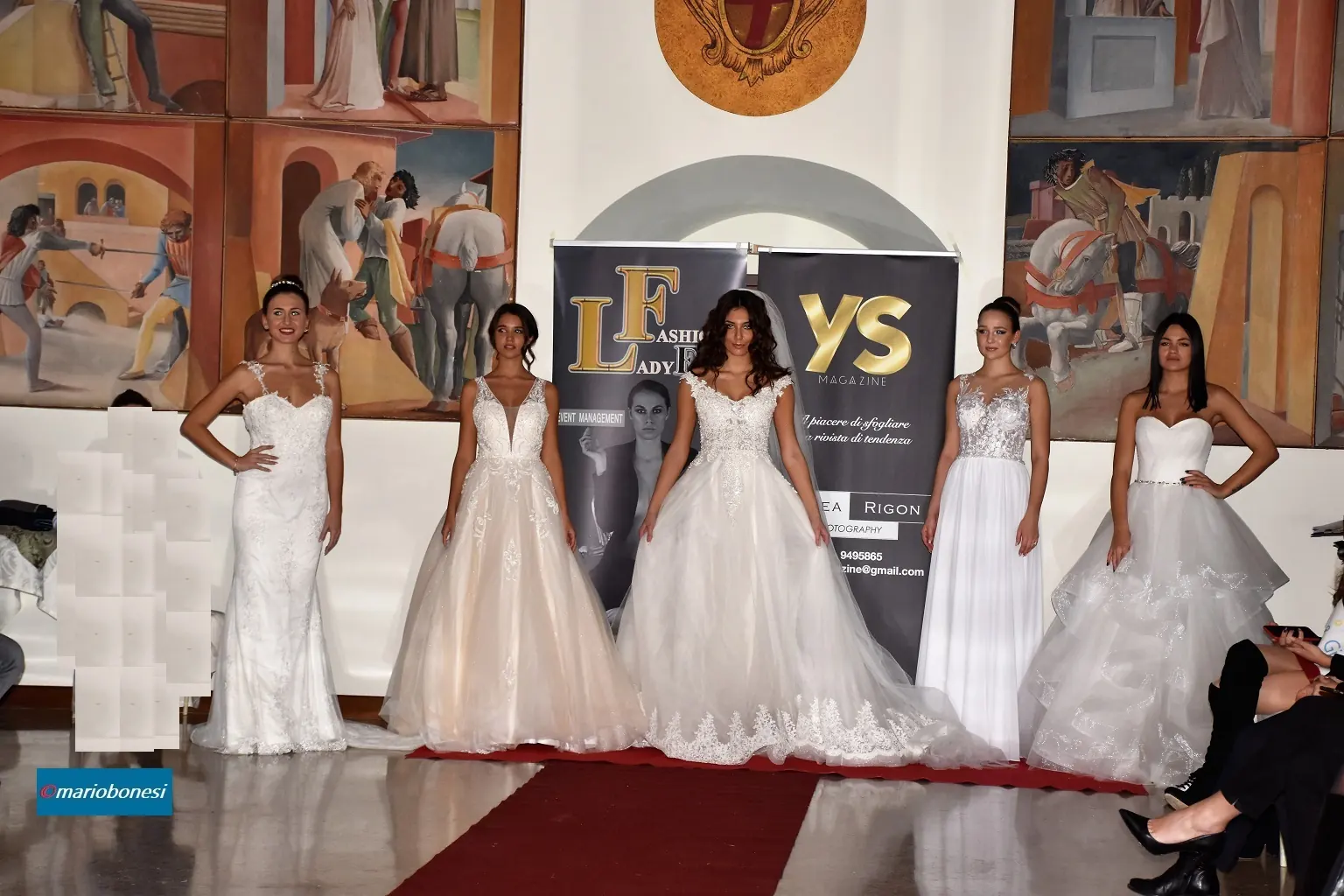 Immagine di una passerella indoor con cinque modelle disposte in fila sul red carpet, tutte indossano abiti da sposa bianchi con stili differenti: gonne in tulle, corpetti in pizzo, linee morbide e dettagli sartoriali. Dietro di loro si trovano due banner con i loghi “LF Fashion Lady Event” e “YS Magazine”. Le pareti della sala sono decorate con grandi affreschi che rappresentano scene storiche con figure in abiti classici. L’illuminazione evidenzia i tessuti chiari e la struttura dei vestiti.