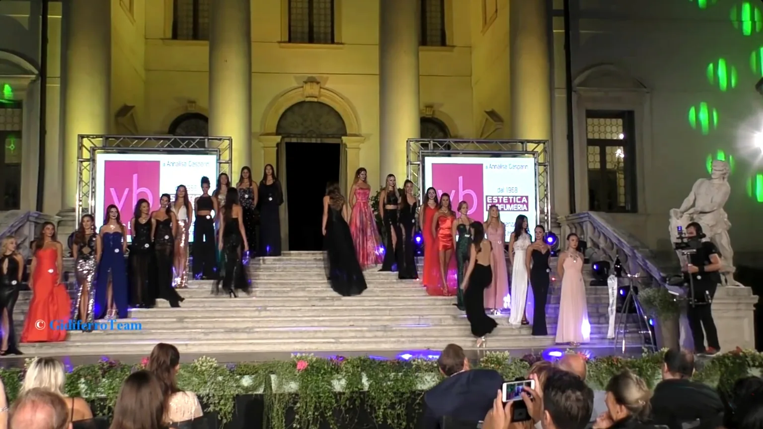 Concorrenti in abito da sera sfilano e posano sui gradini di un edificio storico durante la selezione di Miss Provincia di Vicenza. Le partecipanti indossano abiti eleganti di vari colori e modelli, illuminate da luci da palco. Dietro di loro si vedono due schermi con loghi di sponsor, mentre davanti è presente il pubblico seduto e un operatore video che riprende la scena. L’atmosfera è formale e tipica di un evento di moda all’aperto.