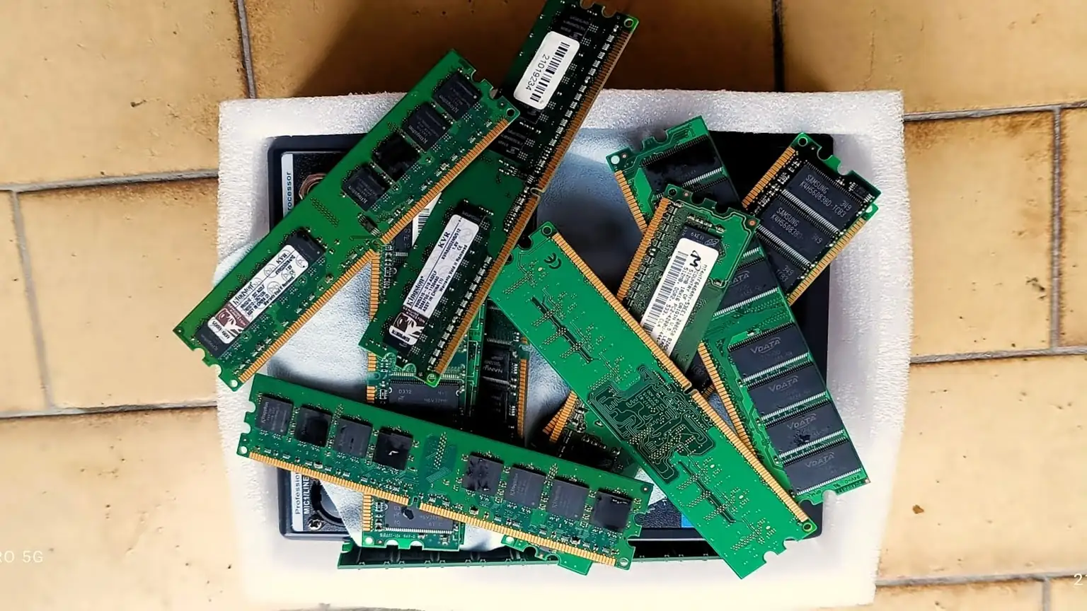 Fotografia che ritrae diversi moduli di memoria RAM appoggiati su un pannello di gommapiuma bianca. I moduli, di colore verde con chip neri e contatti dorati, sono disposti in modo ordinato. Alcuni presentano etichette con informazioni tecniche e numeri di serie. Il supporto poggia su una superficie piastrellata, creando un contesto pulito e ordinato che mette in risalto i componenti elettronici.