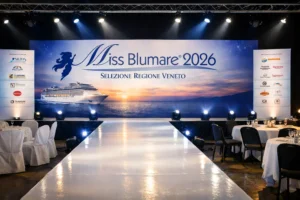 Fotografia panoramica in formato 16:9 che ritrae l’allestimento di una serata dedicata a un concorso di bellezza in una grande sala eventi. In primo piano si vede una passerella lucida bianca che parte dal bordo inferiore dell’immagine e conduce verso un palco sullo sfondo, illuminata da faretti LED disposti lungo i lati. Ai lati della passerella sono presenti file di sedie: a sinistra sedie rivestite da coperture bianche, a destra tavolini rotondi con tovaglie bianche, bicchieri e piccoli centrotavola decorativi. Sul fondo, il palco è dominato da un grande pannello con grafica dedicata al concorso, con scritta “Miss Blumare 2026 – Selezione Regione Veneto”, sagoma stilizzata di una figura femminile e immagine di una nave da crociera su mare calmo al tramonto. Ai lati del palco si trovano pannelli verticali con loghi di sponsor illuminati. L’illuminazione complessiva è fredda e professionale, con toni blu e bianchi che creano un’atmosfera elegante e da evento moda, senza persone riconoscibili in scena.