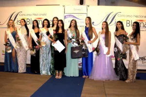 Fotografia orizzontale che mostra le premiate della selezione Miss Blumare Thiene 2026 insieme alla mascotte dell’evento. Le concorrenti, disposte in fila, indossano abiti da sera e fasce identificative, mentre tengono fiori e materiali premio. Davanti a loro è presente la mascotte, riconoscibile per il costume caratteristico e la posizione centrale nella scena. Sullo sfondo compare il banner ufficiale del concorso con loghi e sponsor. L’immagine è luminosa, con messa a fuoco uniforme e dettagli chiaramente leggibili.