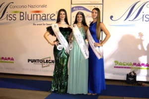 Fotografia orizzontale che mostra le prime tre classificate della selezione Miss Blumare Thiene 2026, in posa sul tappeto blu davanti al backdrop ufficiale. Le concorrenti indossano abiti da sera lunghi di colori diversi e fasce identificative dei rispettivi titoli. La vincitrice Evelyn Priante è al centro con tiara e fascia “Miss Thiene”. Ai lati, le altre due classificate indossano le fasce dei loro sponsor. L’immagine è ben illuminata, con messa a fuoco uniforme e dettagli dei vestiti chiaramente visibili.