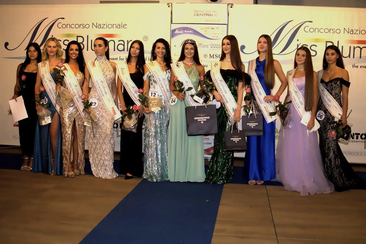 Fotografia orizzontale che ritrae il gruppo completo delle premiate della selezione Miss Blumare Thiene 2026. Undici concorrenti sono disposte in fila sul palco, tutte con abiti da sera lunghi, fasce identificative e bouquet floreali. Alcune tengono in mano sacchetti o attestati. Sullo sfondo è visibile il grande banner del concorso con loghi e sponsor. L’immagine è nitida, con colori naturali e messa a fuoco uniforme su tutto il gruppo.