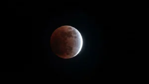 Fotografia della Luna durante la fase avanzata dell’eclissi parziale del 28 agosto 2026, con una copertura di circa l’85%. La superficie lunare appare quasi completamente immersa nell’ombra terrestre, assumendo una tonalità rosso‑bruna tipica delle eclissi, mentre una sottile falce luminosa è visibile sul bordo destro. Il cielo è nero e mette in risalto il contrasto tra la parte oscurata e la piccola porzione ancora illuminata. L’immagine documenta la fase prossima al massimo dell’eclissi.