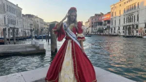Fotografia scattata lungo un canale veneziano. Una ragazza indossa un abito storico rosso con ricami dorati, maniche ampie e dettagli bianchi, insieme a una fascia cerimoniale e un copricapo decorato. Sullo sfondo si vedono edifici veneziani con archi, balconi e finestre, oltre a barche ormeggiate lungo il canale. L’immagine documenta un momento di rappresentanza in abito tradizionale nel contesto urbano di Venezia.