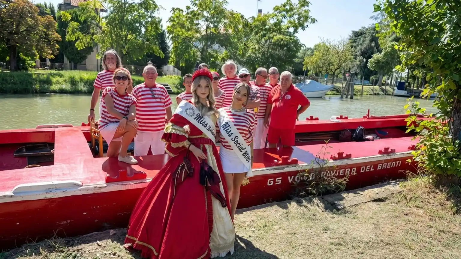 Fotografia scattata lungo un canale, dove un gruppo di vogatori posa accanto a una barca rossa con la scritta GS VOGA Riviera del Brenta. La maggior parte dei ragazzi indossa maglie a righe rosse e bianche, mentre davanti al gruppo si trovano due ragazze: una con abito storico rosso e oro e fascia Miss Riviera del Brenta, l’altra con abbigliamento bianco e fascia Miss Social RDB. Sullo sfondo si vedono alberi, prato e il corso d’acqua, in un contesto di evento tradizionale all’aperto.