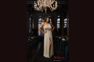 La patron Elisa Bagordo, presentatrice e ideatrice del concorso, è ritratta al centro del palco durante la finale di Miss Venice Beach 2022, indossando un abito bianco monospalla mentre introduce le concorrenti. Le ragazze, disposte in fila, indossano abiti blu con logo dorato. La sala è elegante, con lampadari, tende e pubblico seduto. L’immagine testimonia la continuità del format che ha portato alla storica edizione 2026 e alla vittoria di Maddalena Rossi.