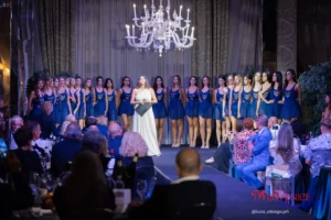 La patron Elisa Bagordo durante una serata ufficiale del concorso Miss Venice Beach, ritratta al centro del palco mentre indossa un abito bianco monospalla e tiene in mano una cartellina. Davanti a lei è disposta una fila di concorrenti in abiti blu con logo dorato, tutte in posizione frontale. La sala è elegante, con un grande lampadario bianco sospeso, tende alte e tavoli apparecchiati dove il pubblico osserva la scena. Sullo sfondo sono presenti elementi decorativi e luci che valorizzano l’atmosfera formale dell’evento. L’immagine documenta il ruolo centrale della patron nella conduzione del concorso e nella finale che ha portato Maddalena Rossi alla vittoria.
