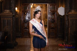 Maddalena Rossi, vincitrice di Miss Venice Beach 2026, ritratta in un ambiente storico veneziano con pannelli lignei decorati. Indossa un abito blu, la corona ufficiale e la fascia della vincitrice, mentre tiene in mano il trofeo in vetro. Sullo sfondo si intravede una sala illuminata e un pannello fotografico professionale. L’immagine rappresenta il momento celebrativo della sua incoronazione, simbolo della “magia dal Lido alla corona”.