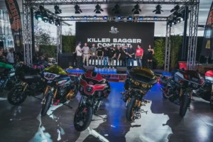 Fotografia scattata all’interno di un padiglione del Moto Show, con diverse moto bagger personalizzate esposte in primo piano. Le motociclette presentano verniciature elaborate, carene modificate e dettagli cromati. Sul palco, in fondo alla scena, è visibile un gruppo di persone davanti a un grande schermo nero con la scritta “Eternal City – Killer Bagger Contest”. L’illuminazione professionale e la struttura metallica del soffitto indicano un evento organizzato, con pubblico e partecipanti presenti nell’area.