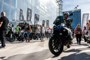 Fotografia di un’area esterna del Moto Show, con un edificio moderno in vetro sullo sfondo che riporta la scritta “Eternal City Moto Show 27-28 Settembre”. Lungo il percorso sono posizionate bandiere nere con il logo BMW Motorrad. Diversi motocicli sono parcheggiati in fila, mentre visitatori camminano osservando i modelli esposti. In primo piano, un motociclista con casco e abbigliamento tecnico è seduto su una moto BMW di colore blu. L’atmosfera è vivace e rappresenta una tipica area test ride o esposizione all’aperto.