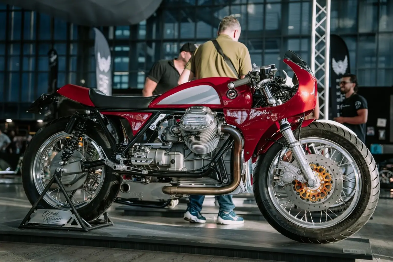 Fotografia di una moto custom esposta all’interno di un salone motociclistico. Il veicolo presenta una livrea rossa e argento, componenti in metallo lucidato, un grande motore a vista e una carenatura in stile café racer. La moto è posizionata su un supporto con la scritta “Moto Parigiuse”. Sullo sfondo si vedono persone che osservano l’esposizione e pannelli in vetro che suggeriscono un ambiente moderno e luminoso. L’immagine documenta un modello artigianale presentato durante l’evento.