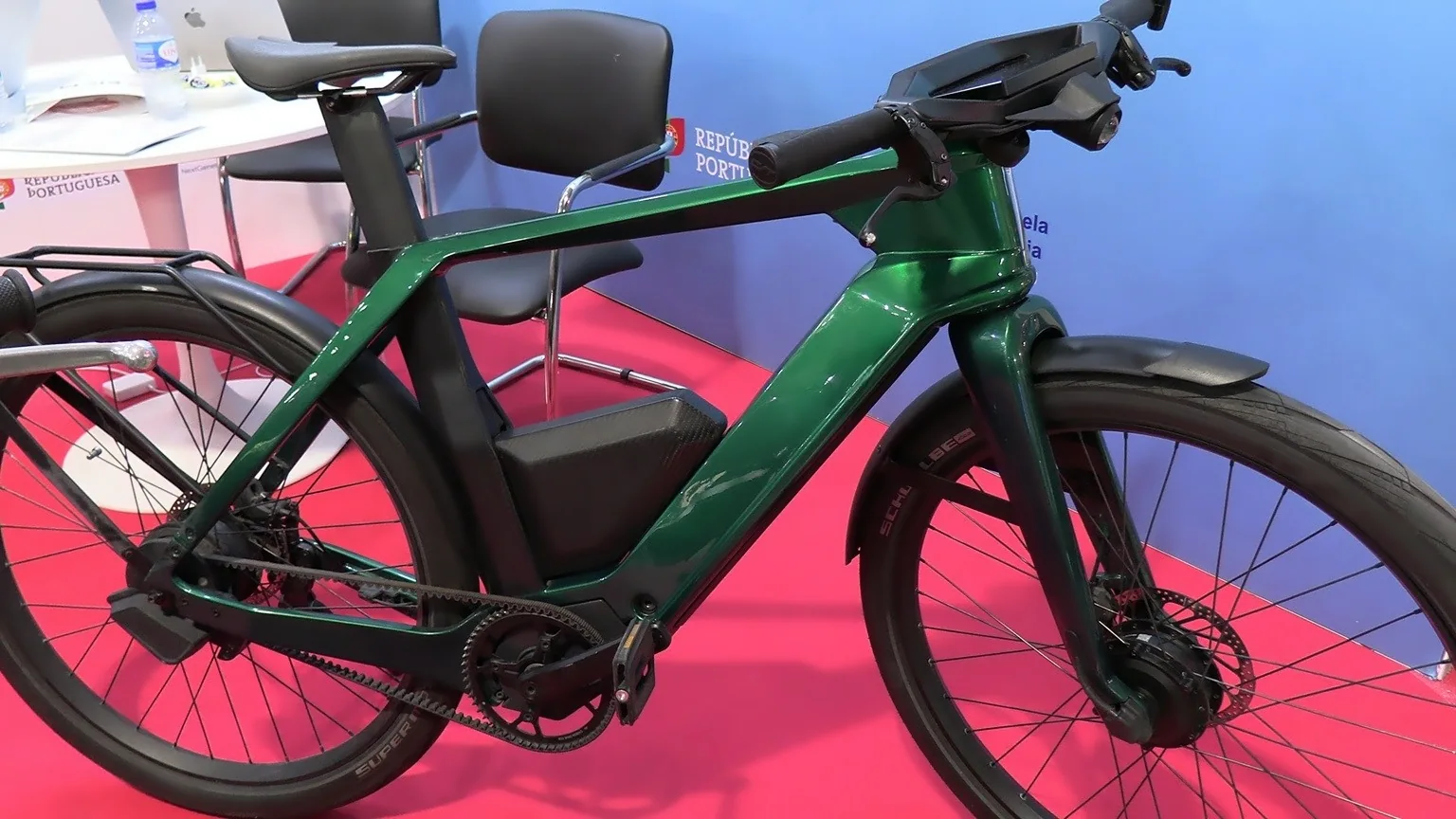 Fotografia scattata in uno spazio espositivo interno che mostra una bicicletta elettrica dal design moderno, posizionata su pavimento rosso. Il telaio è angolare e di colore verde metallizzato, con batteria integrata nel tubo obliquo e trasmissione a cinghia. Le ruote montano freni a disco e pneumatici con marchio visibile. Il manubrio ha comandi integrati e linee aerodinamiche. Sullo sfondo si vedono sedie, un tavolo con laptop e una parete blu con scritte istituzionali. L’immagine documenta un modello di e‑bike presentato durante un evento dedicato alla mobilità.