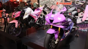 Immagine scattata in un’area espositiva interna con due motociclette custom decorate con grafiche ispirate al personaggio Kuromi. La moto a sinistra è rosa con dettagli viola e motivi a scacchi sul parafango, mentre quella a destra è viola con elementi neri e rosa e la scritta “Kuromi” sulla carenatura. Sullo sfondo è presente un grande pannello con illustrazioni del personaggio e la dicitura “Kuromi 20th Anniversary”. Alcune persone osservano le moto. La scena documenta un’esposizione tematica all’interno di un evento motociclistico.