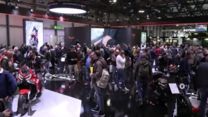 Fotografia scattata in un padiglione fieristico affollato, dove numerosi visitatori osservano motociclette esposte su pavimento nero lucido. In alto è visibile un grande pannello con la scritta “Moto Morini”, accompagnato da schermi digitali che mostrano video promozionali, tra cui un’immagine di un motociclista su una moto adventure rossa. Attorno allo stand si notano altri marchi e strutture espositive. L’immagine documenta l’atmosfera di una fiera motociclistica con forte affluenza di pubblico.