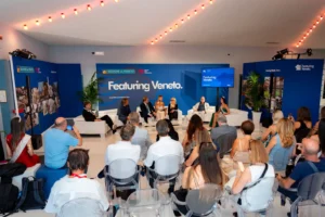 Fotografia di un panel ufficiale nello spazio “Featuring Veneto” della Regione del Veneto e Veneto Film Commission: relatori seduti su divani bianchi dialogano davanti a un pubblico disposto su sedie trasparenti. Sullo sfondo, un grande pannello blu con loghi istituzionali e la scritta “Featuring Veneto”. L’ambiente è moderno, illuminato da luci decorative e arricchito da piante ornamentali. L’immagine documenta il momento di presentazione del progetto legato alle Città Murate.