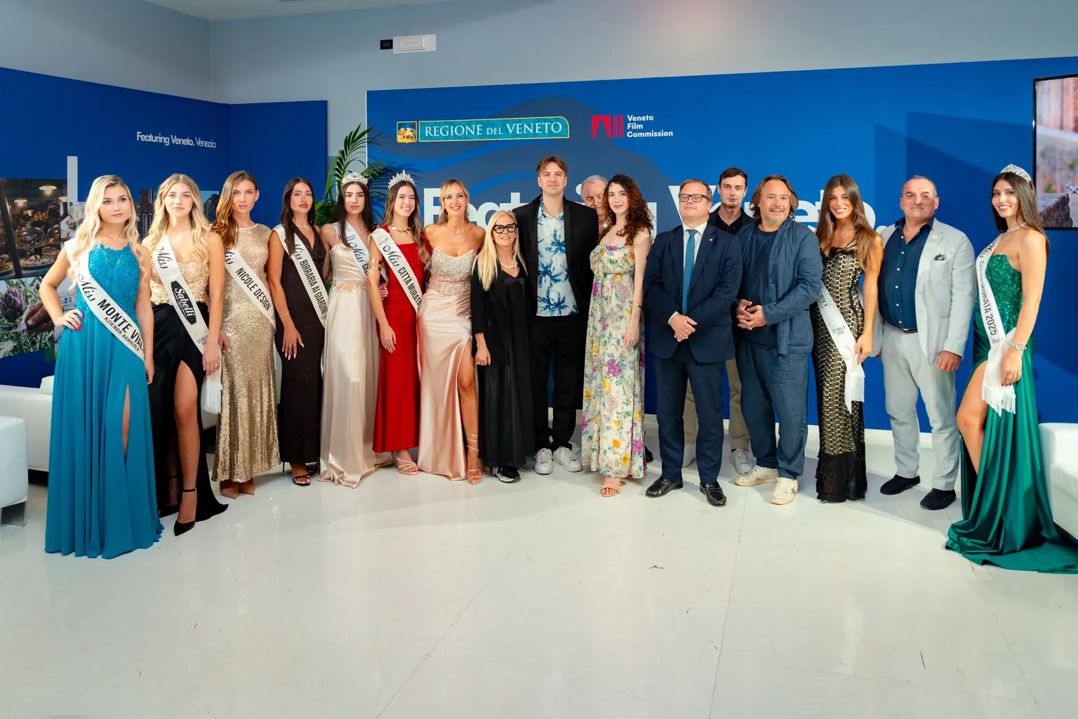 Fotografia di gruppo con Miss in abito da sera e fasce ufficiali, affiancate da rappresentanti istituzionali e ospiti in abiti formali. Sullo sfondo, il pannello blu della Regione del Veneto e della Veneto Film Commission. L’immagine documenta il momento di incontro tra le Miss Città Murata e le figure istituzionali presenti alla Mostra del Cinema, evidenziando il ruolo del concorso nella promozione culturale del territorio.