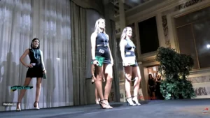 Fotografia scattata durante la finale di Miss Venice Beach 2024. Sul palco, tre concorrenti indossano abiti neri corti, collane lunghe e décolleté con tacco alto. Ognuna porta una piccola borsa verde e una targhetta numerata: sono visibili i numeri 19 e 21. Le partecipanti posano in linea davanti alle tende del palco, illuminate da luci sceniche. Sullo sfondo si intravedono pareti con decorazioni marmoree e alcune persone vicino a un ingresso laterale. L’immagine documenta un momento della sfilata serale, con le concorrenti impegnate nella presentazione ufficiale sul palco.