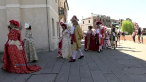  Fotografia di un gruppo di figuranti che cammina lungo una strada della Riviera del Brenta indossando abiti storici del Settecento: le donne portano vestiti riccamente decorati con trame floreali, pizzi e gonne ampie, mentre gli uomini indossano giacche lunghe, camicie con jabot, calzoni al ginocchio, calze bianche e cappelli tricorni con piume. Sullo sfondo si vedono edifici chiari con balconi e finestre, alcuni spettatori che osservano e scattano foto, e un’atmosfera di rievocazione storica tipica di Riviera Fiorita. L’immagine documenta il passaggio dei figuranti durante il corteo acqueo e le attività collaterali della manifestazione.