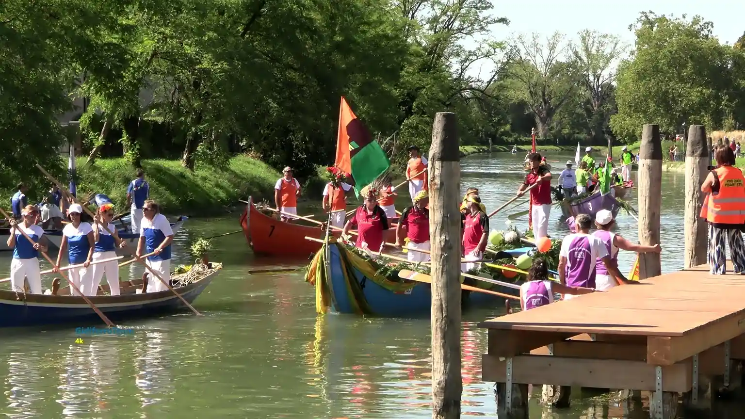 Fotografia del corteo acqueo di Riviera Fiorita sul Naviglio Brenta: diverse imbarcazioni in legno avanzano sul fiume, ciascuna con equipaggi in abbigliamento coordinato nei colori blu, rosso, arancione, viola e verde. Alcune barche sono decorate con fronde, bandiere e elementi ornamentali tipici della manifestazione. Ai margini del fiume si nota un pontile in legno con persone che osservano o assistono i vogatori, mentre la vegetazione verde e fitta crea una cornice naturale attorno alla scena. L’immagine rappresenta uno dei momenti più caratteristici della tradizione remiera della Riviera del Brenta.