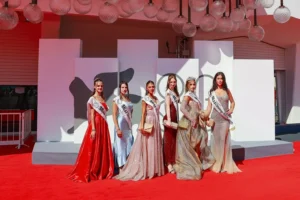 immagine orizzontale scattata su un red carpet, con sei giovani donne in abiti da sera eleganti, disposte in fila davanti a un fondale bianco con logo nero stilizzato. Ognuna indossa una fascia con il proprio titolo, e una di loro porta una corona, segno della vittoria nel concorso. Le ragazze posano sorridenti, illuminate dalle luci dell’evento, in un contesto formale che richiama la Mostra del Cinema di Venezia. L’atmosfera è professionale e celebrativa, con attenzione alla moda, alla presentazione e alla rappresentazione ufficiale delle Miss Riviera del Brenta.