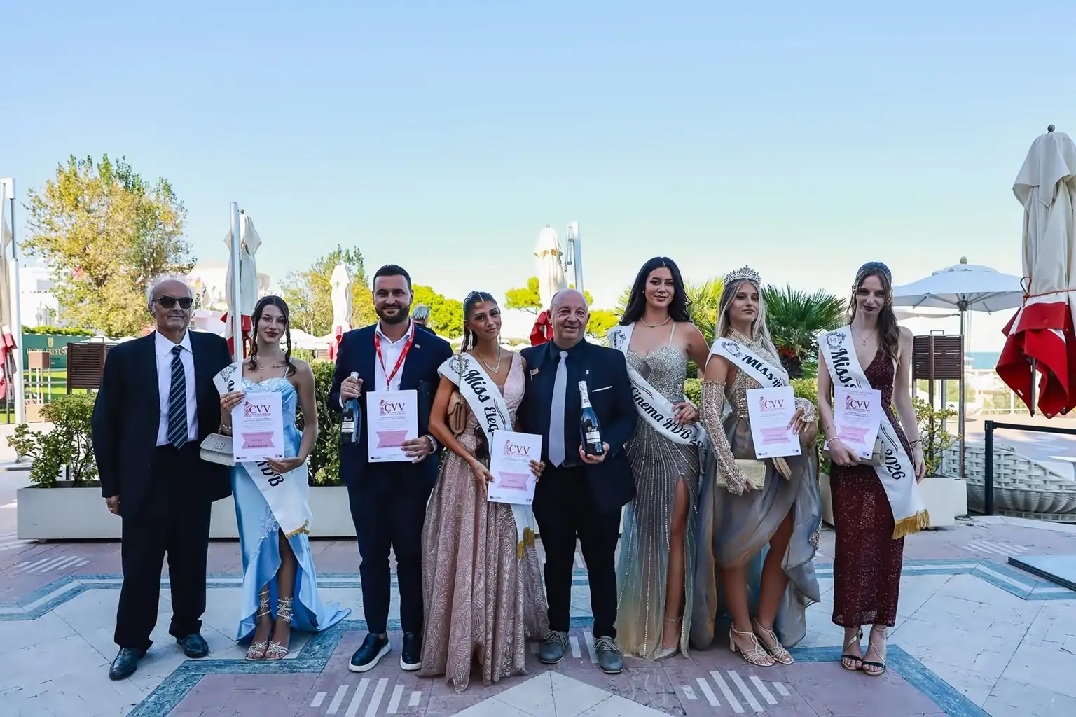 immagine orizzontale scattata all’aperto su una terrazza con pavimento in pietra, ombrelloni chiari e vegetazione sullo sfondo. Otto persone posano in gruppo: alcune indossano abiti da sera, altre completi eleganti. Diverse ragazze portano fasce con titoli come “Miss Eleganza”, “Miss Cinema” e “Miss CVV 2026”, e una di loro indossa una corona. Alcuni partecipanti tengono in mano certificati con la sigla “CVV” e bottiglie premio. L’atmosfera è festosa e istituzionale, tipica di una cerimonia di premiazione legata al cinema veneto e alle attività del concorso Miss Riviera del Brenta.
