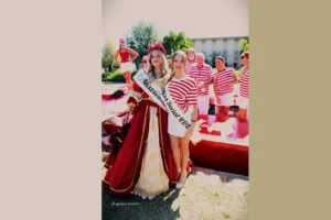  immagine orizzontale scattata all’aperto durante una manifestazione remiera. In primo piano due giovani donne: una indossa un abito storico rosso e oro con gonna ampia e una fascia “Miss Riviera del Brenta 2018”, l’altra porta una maglia a righe rosse e bianche, una gonna bianca e la fascia “Miss Social RDB”. Sullo sfondo si vedono altre persone con maglie a righe coordinate e strutture rosse legate alla voga. La scena è luminosa, con cielo sereno, e rappresenta un momento della tradizione veneziana, collegato alla partecipazione delle Miss Riviera del Brenta alla Regata Storica. In basso è visibile la firma del fotografo “ph giuliano marcato”.