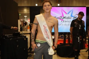 Inquadratura in piano americano di Nicolas Camplani, vincitore del titolo di Mister Rombo di Tuono 2026. Il giovane bresciano posa a torso nudo indossando la fascia bianca e rossa 'Mister Rombo di Tuono Finalista' a tracolla, pantaloncini grigi sportivi con numero di gara e tatuaggi visibili sulle braccia e sul petto.