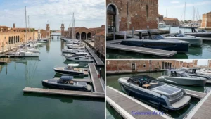 Veduta panoramica ad ampio raggio del bacino dell'Arsenale di Venezia, con numerose imbarcazioni a motore e a vela ormeggiate lungo i pontili galleggianti. Sullo sfondo si ergono le storiche torri dell'Arsenale e le antiche tese in mattoni a vista sotto un cielo leggermente nuvoloso.