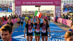 Un'inquadratura ravvicinata e frontale in piena luce solare cattura tre giovani donne runner, sorridenti ed entusiaste, che festeggiano appena dopo aver tagliato il traguardo della 40ª Wizz Air Venicemarathon. Indossano magliette tecniche nere identiche con la scritta fucsia e bianca "40ª WIZZ AIR VENICEMARATHON" e i loro numeri di pettorale: la donna a sinistra (numero 4001, italiana), al centro (numero 4002), e a destra (numero 4003). La donna al centro tiene alta una bandiera italiana sventolante. Hanno tutte la medaglia da finisher al collo e si abbracciano con gioia, guardando la bandiera. Si trovano sulla pista d'arrivo blu patterned, con una folla densa e sfocata di altri corridori e spettatori dietro di loro. Sopra di loro, una grande struttura di traguardo in acciaio e banner rosa/viola con la scritta "40ª WIZZ AIR VENICEMARATHON" e vari sponsor, tra cui Joma, Hyundai e Wizz Air, e un timer digitale (sfocato, circa 12:07:07). Sullo sfondo, oltre l'acqua della laguna con barche, si staglia il campanile di San Marco e il Palazzo Ducale di Venezia sotto un cielo sereno.