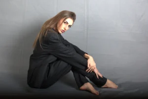 Ritratto in studio di Giulia Urbinati, seduta a terra in posa raccolta su uno sfondo grigio uniforme. Indossa un completo sartoriale nero, composto da una giacca oversize e pantaloni coordinati, ed è a piedi nudi. I suoi lunghi capelli castani le scendono morbidi sulle spalle e il suo sguardo è rivolto intensamente verso l'obiettivo. La posa è introspettiva, con le mani che le cingono le ginocchia piegate. L'illuminazione morbida e diffusa mette in risalto la texture del tessuto e la profondità del suo sguardo, creando un'atmosfera elegante e minimalista. L'inquadratura cattura la figura intera.