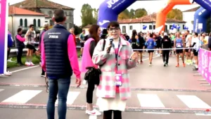 La presentatrice Beatrice Tomada con occhiali e una giacca a quadri rosa e verde è in piedi al centro, tenendo un microfono e parlando. Dietro di lei ci sono un uomo con una giacca a vento Joma e una donna con un giubbotto rosa. Sullo sfondo si vede un arco blu gonfiabile con il logo Joma e un arco arancione gonfiabile, mentre numerosi podisti attraversano la linea di partenza della maratona. Degli edifici sono visibili sullo sfondo.