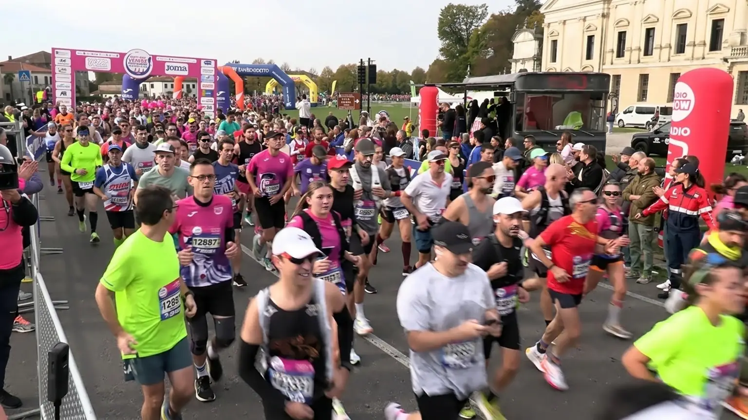 Un'ampia visuale della linea di partenza della Venice Marathon con una folla densa di podisti vestiti con abiti sportivi colorati. Al centro dell'immagine, in primo piano, un uomo indossa una maglietta verde brillante. Sulla sinistra, un podista indossa una maglietta rosa. Sullo sfondo ci sono due grandi archi gonfiabili: uno rosa con loghi Joma e uno blu con un logo. Sulla destra, è visibile un camion e una struttura di Radio Company gonfiabile rossa. Molte persone si accalcano ai lati della strada per guardare la partenza.