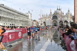 Atleti della Venicemarathon attraversano Piazza San Marco nel centro storico di Venezia durante uno dei momenti più iconici della maratona. Sullo sfondo si distingue la Basilica di San Marco, mentre lungo il percorso il pubblico assiste e applaude i runner. Il selciato riflette l'immagine dei partecipanti e dei monumenti veneziani, creando uno degli scorci più suggestivi della manifestazione.