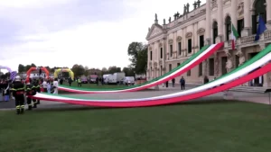Immagine a campo largo scattata nel parco antistante Villa Pisani a Stra. Alcuni operatori dei Vigili del Fuoco e volontari in divisa tengono teso un lunghissimo nastro con i colori della bandiera italiana (verde, bianco e rosso) che si estende per decine di metri sul prato verde, sullo sfondo della storica villa veneta e dei gonfiabili dell'area di partenza.