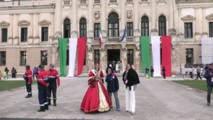 Inquadratura dal basso di fronte alla facciata monumentale di Villa Pisani. Al centro, la Miss Riviera del Brenta indossa un ampio abito tradizionale rosso e oro con una fascia elegante, affiancata da due donne con microfono che conducono le interviste. Sulla sinistra, alcuni operatori di sicurezza in divisa osservano la scena, mentre a destra si intravedono spettatori e biciclette.