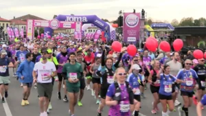 Fotografia in primo piano della folla di podisti in festa subito dopo il via della maratona. Un gruppo di atleti sorridenti avanza lungo la strada asfaltata indossando pettorali di gara, abbigliamento tecnico e zainetti da running; alcuni partecipanti tengono in mano dei palloncini rossi, mentre sullo sfondo spiccano i grandi archi gonfiabili viola e le strutture della manifestazione.
