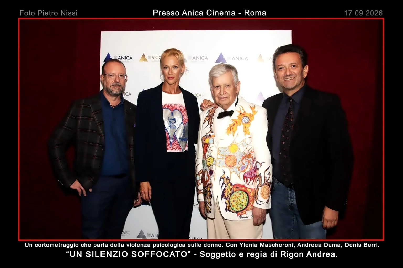 Foto di gruppo realizzata presso il Cinema Anica di Roma durante la presentazione del cortometraggio Un Silenzio Soffocato. Quattro persone posano davanti al pannello ufficiale dell'evento con i loghi ANICA. Tra i presenti figurano il regista Andrea Rigon e alcuni protagonisti e sostenitori del progetto cinematografico dedicato al tema della violenza psicologica sulle donne. Sullo sfondo è visibile il backdrop istituzionale dell'Anica, mentre in primo piano spicca un elegante completo bianco decorato artisticamente indossato da uno degli ospiti. La fotografia documenta uno dei momenti ufficiali della serata romana dedicata alla promozione del cortometraggio presentato il 17 settembre 2026.